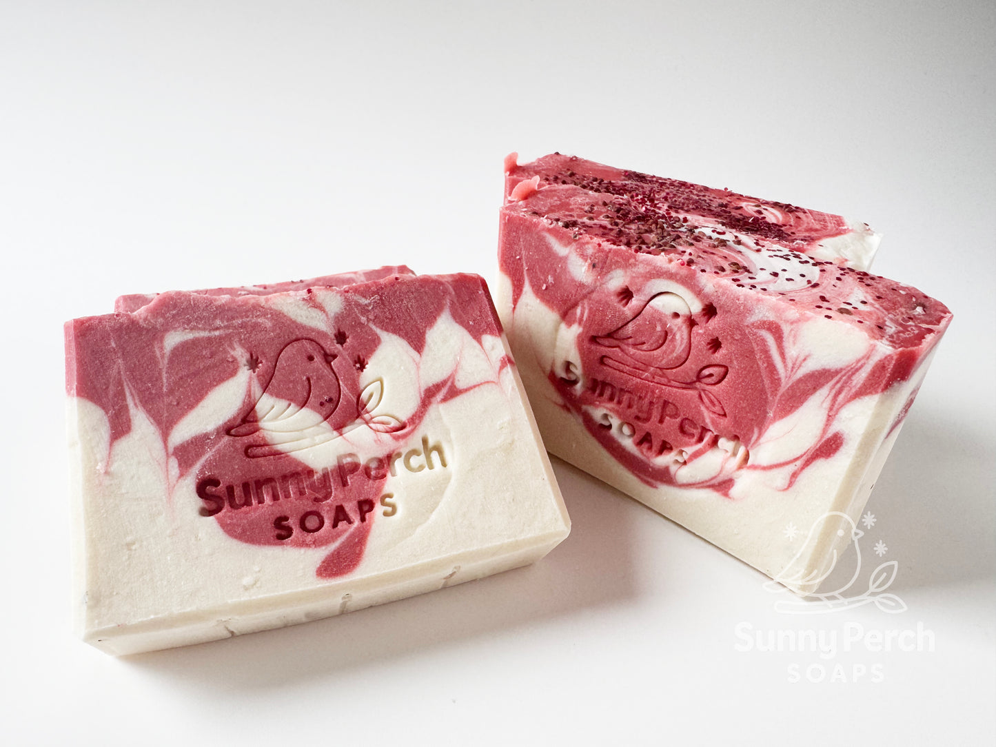 Four bars of soap with Sunny Perch Soaps brand stamp on white background. Soap is mainly white in color with red swirling. Top of soap bar is decorated with cranberry seeds and biodegradable glitter.