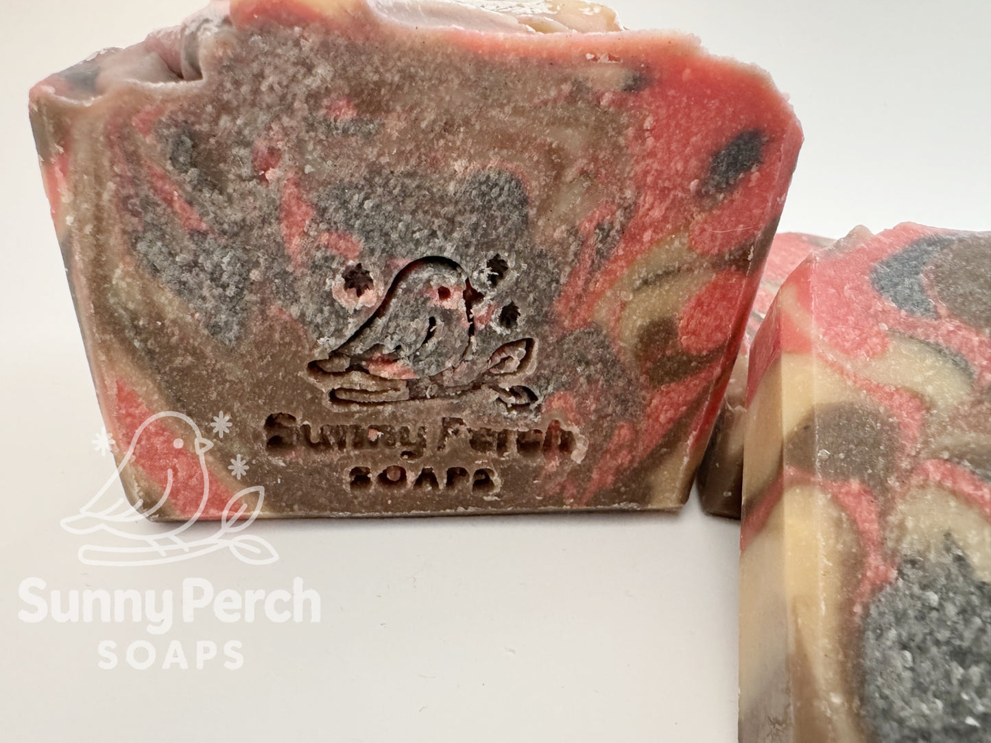 Close up of one bar of marbled soap with 'Sunny Perch Soaps' branding on a white background. Soap color is light caramel with dark tan and red swirling colors.