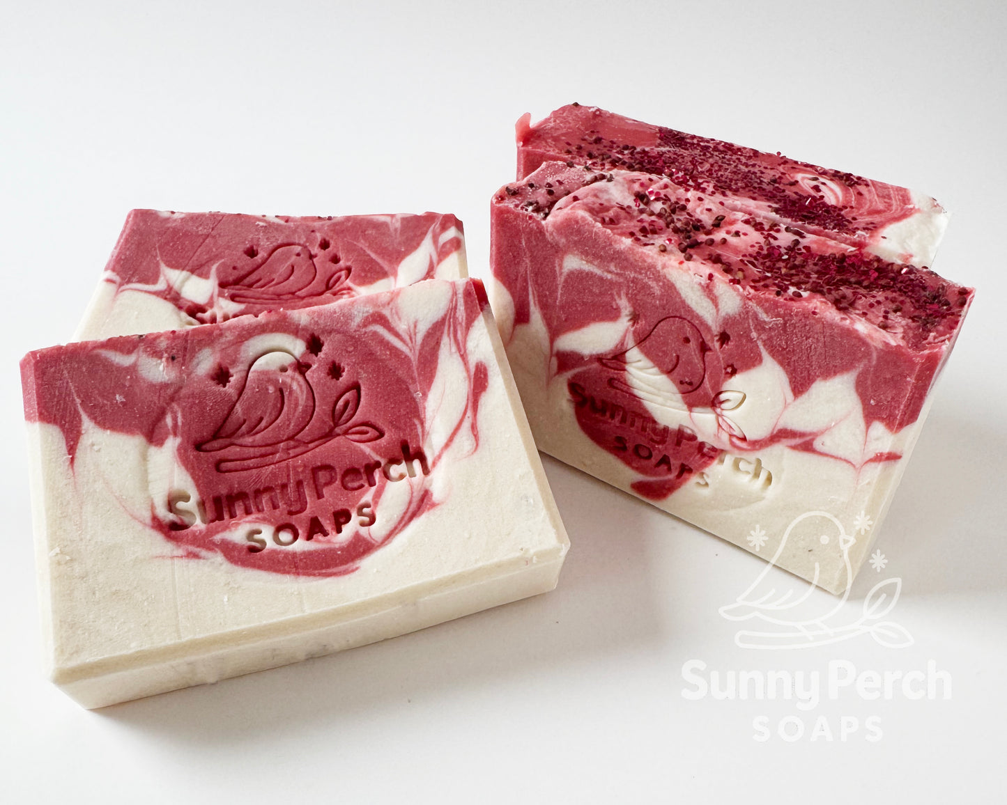Four bars of soap with Sunny Perch Soaps brand stamp on white background. Soap is mainly white in color with red swirling. Top of soap bar is decorated with cranberry seeds and biodegradable glitter.