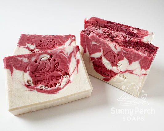 Four bars of soap with Sunny Perch Soaps brand stamp on white background. Soap is mainly white in color with red swirling. Top of soap bar is decorated with cranberry seeds and biodegradable glitter.
