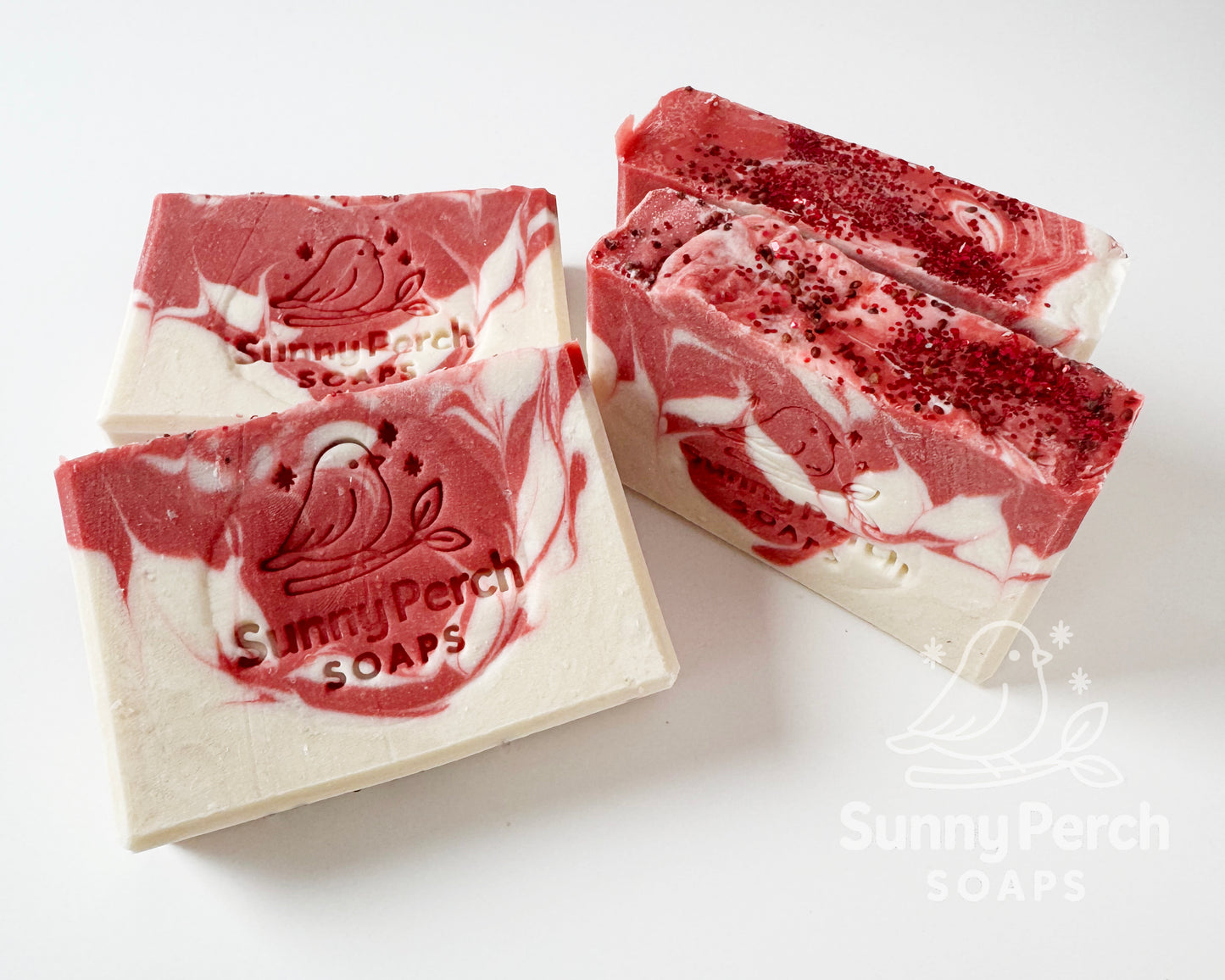 Four bars of soap with Sunny Perch Soaps brand stamp on white background. Soap is mainly white in color with red swirling. Top of soap bar is decorated with cranberry seeds and biodegradable glitter.