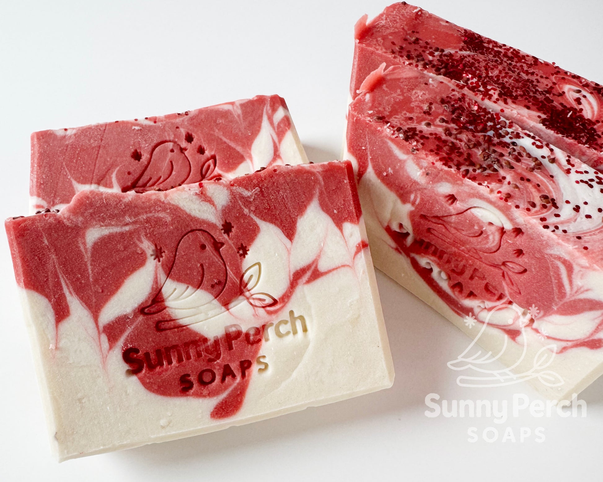 Closer image of four bars of soap with Sunny Perch Soaps brand stamp on white background. Soap is mainly white in color with red swirling. Top of soap bar is decorated with cranberry seeds and biodegradable glitter.