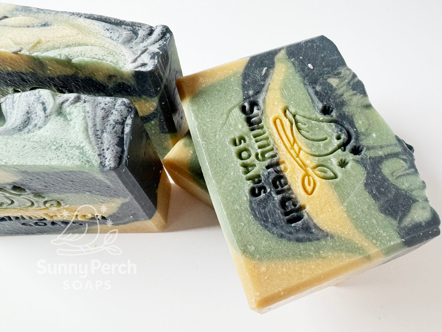 Closer image of four soap bars with a visible brand logo on a white background. Soap color is swirls of green, dark gray and yellow in color.