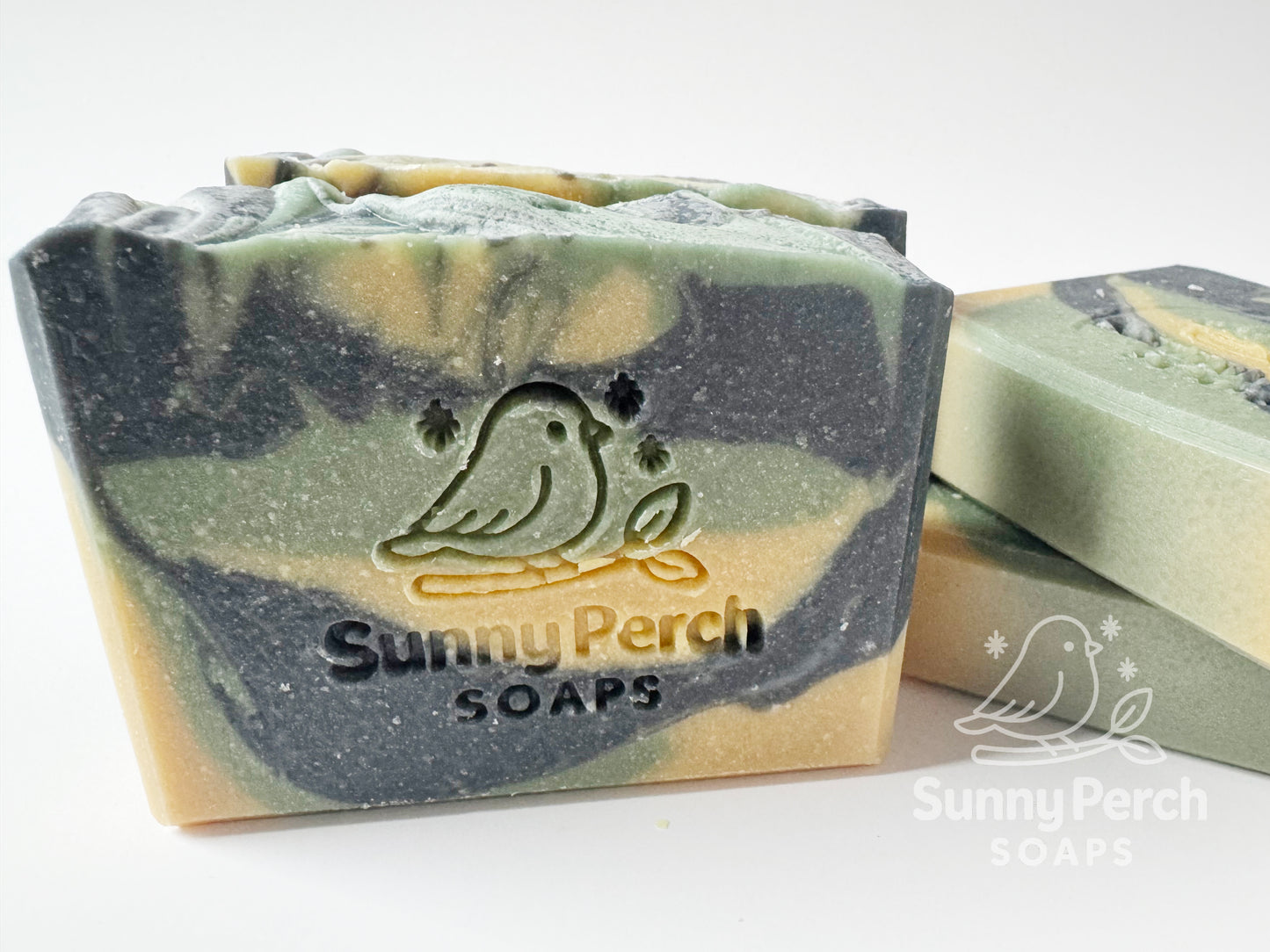 Closer image of four soap bars with a visible brand logo on a white background. Soap color is swirls of green, dark gray and yellow in color.