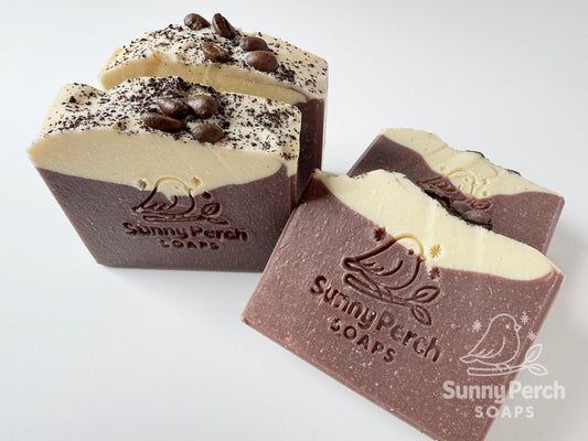Three bars of soap with visible branding on a white background. Soaps are mainly brown in color with cream on top. Decorated with coffee beans and coffee grounds.